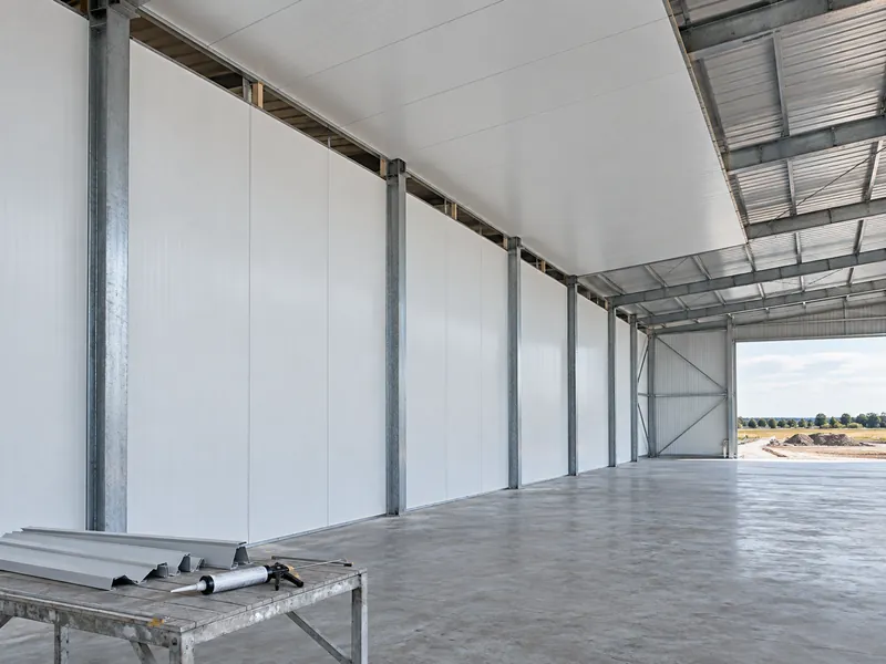 Intérieur d'un entrepôt frigorifique pendant la pose des panneaux, avec des panneaux sandwich isolants fixés à la charpente métallique et des joints mur-toiture encore ouverts en attente d'étanchéité